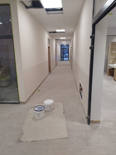 Długi korytarz w budynku po świeżym pomalowaniu ścian na dwukolorowo, widoczne dwa wiadra z farbą na kartonie zabezpieczającym podłogę.