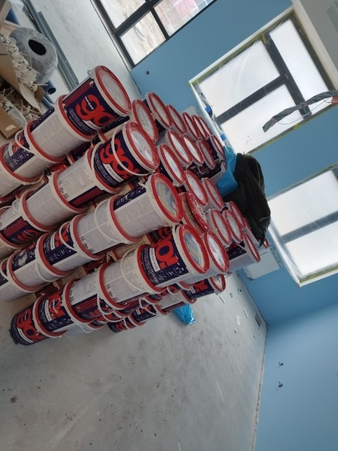 Stos kilkudziesięciu wiader z farbą marki Q4 ułożonych w piramidę na podłodze w pomieszczeniu z niebieskimi ścianami i oknami zabezpieczonymi folią malarską.