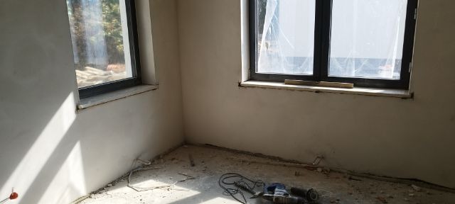 Pokój w trakcie remontu; widoczne dwa okna, ściany po szpachlowaniu, przygotowane do dalszych prac wykończeniowych, na podłodze narzędzia budowlane i resztki materiałów.