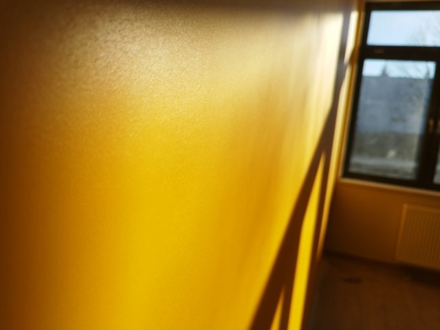 Ściana pomalowana na intensywny żółty kolor, oświetlona popołudniowym słońcem, z wyraźnym cieniem ramy okiennej, widoczne okno i fragment grzejnika w tle.