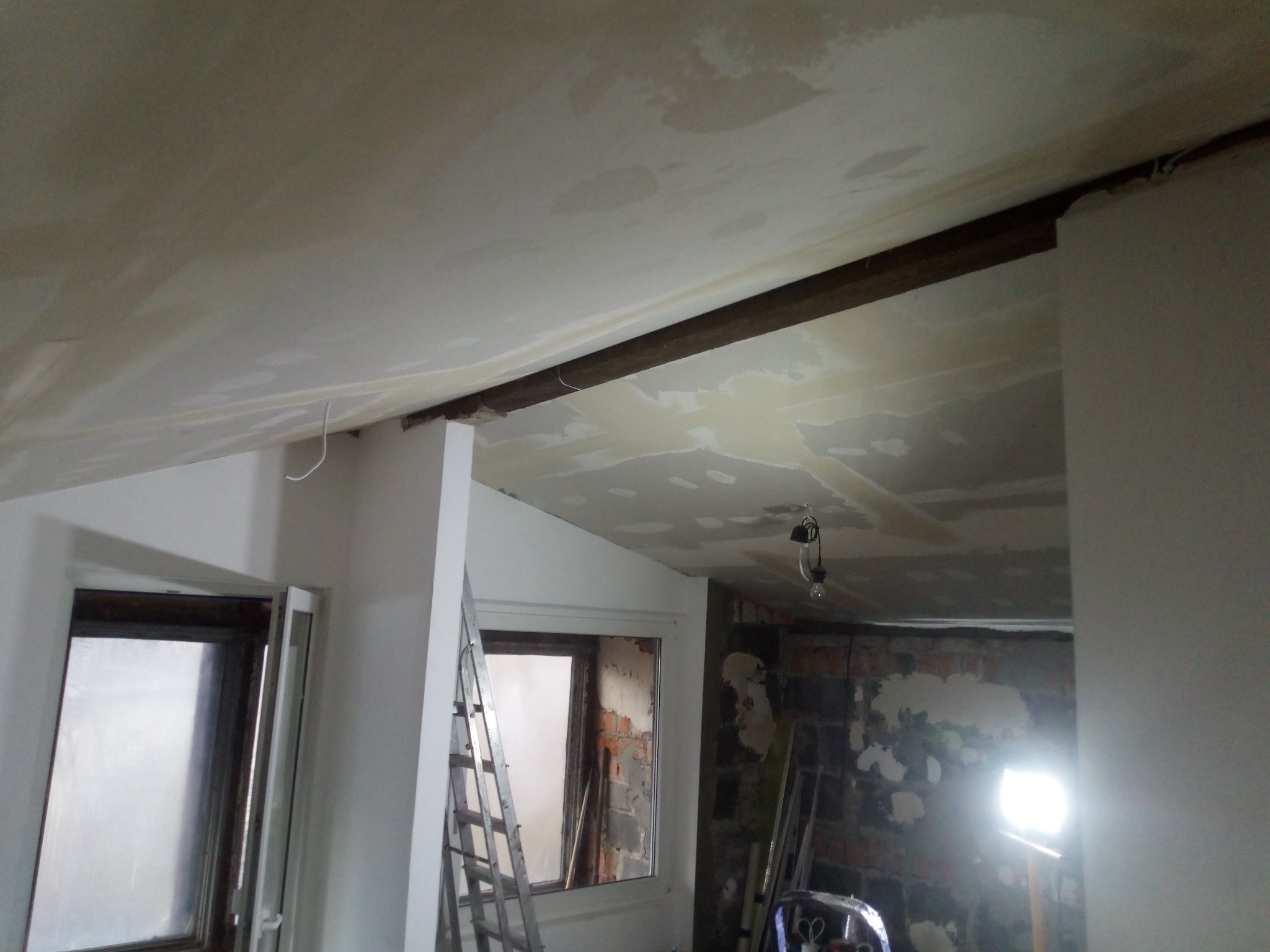 Niedokończony sufit z widocznymi łatami szpachli, fragmentami odsłoniętej cegły i drewnianą belką, drabina oparta o okno w tle.