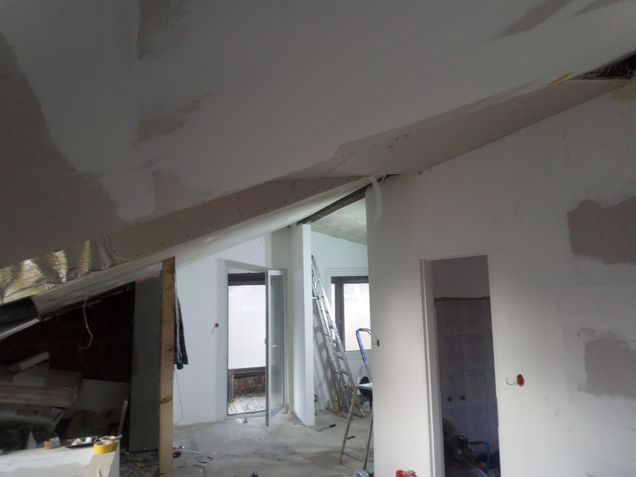 Poddasze w trakcie prac wykończeniowych: widoczne ocieplenie, płyty kartonowo-gipsowe na skosach sufitu oraz ściany po wstępnym szpachlowaniu, aluminiowa drabina w tle.