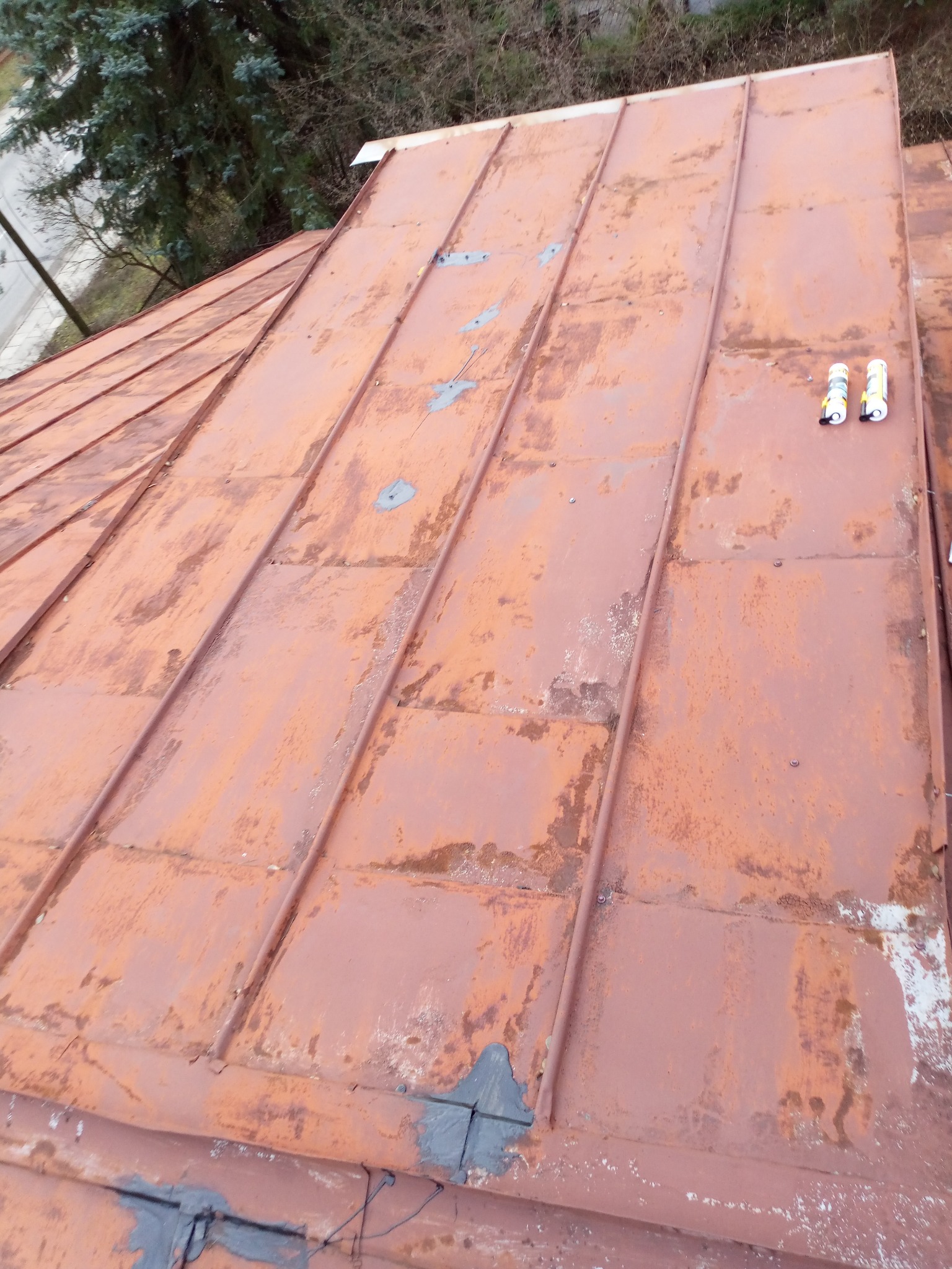 Zardzewiały metalowy dach z widocznymi łatami i dwoma tubami uszczelniacza na górze, tło stanowią drzewa.