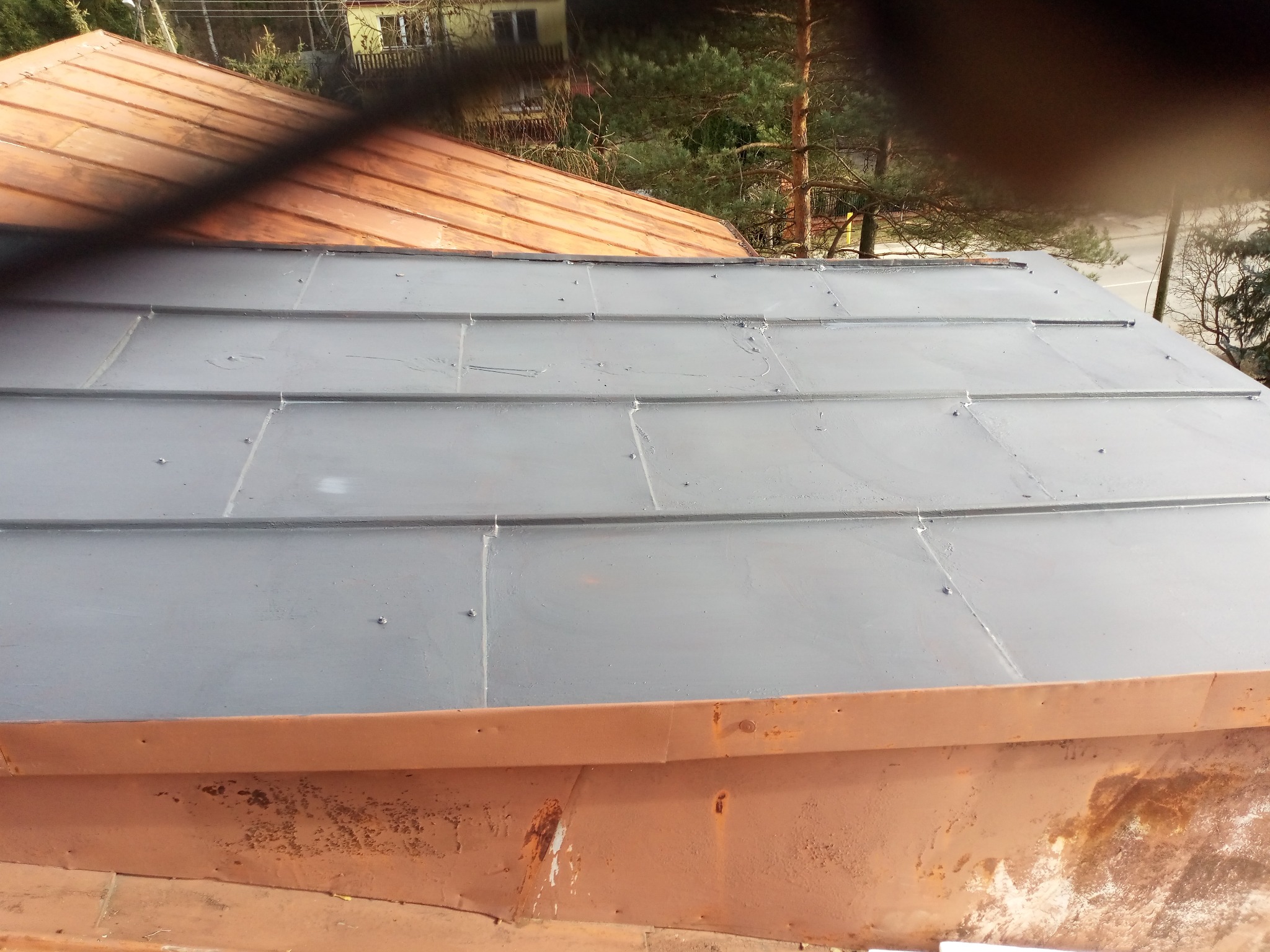 Szare, metalowe panele dachowe, częściowo pomalowane, z widocznymi śrubami mocującymi, sąsiadujące z miedzianym dachem i elementami orynnowania w kolorze rdzy.