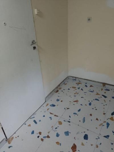 Pusta przestrzeń w rogu pokoju z białymi ścianami w odcieniach beżu, białe drzwi po lewej stronie i podłoga wyłożona płytkami w mozaikę z niebieskimi, brązowymi i pomarańczowymi elementami.