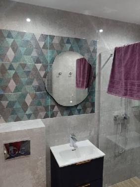 Wykończona łazienka z geometrycznymi płytkami w odcieniach szarości, zieleni i błękitu, okrągłe lustro z oświetleniem LED, biała umywalka na czarnej szafce, szklana kabina prysznicowa z widocznym...