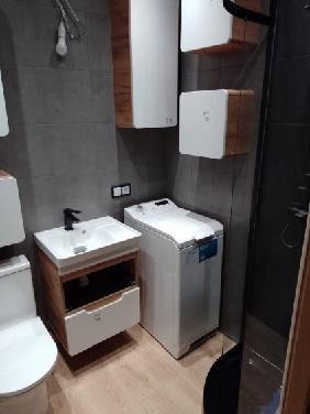 Nowoczesna łazienka z szarą płytką, białą pralką i umywalką z drewnianą szafką. Widoczne wiszące białe szafki z elementami drewna. Minimalistyczny wystrój, surowy styl.