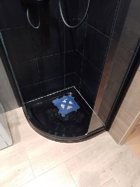 Ciemna kabina prysznicowa z brodzikiem po remoncie, widoczna niebieska mata antypoślizgowa i jasna drewniana podłoga.