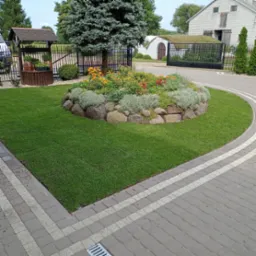 Ogród z równo przystrzyżonym trawnikiem, okrągłą rabatą kwiatową otoczoną kamieniami, studnią z drewnianą obudową i budynkiem z dachem pokrytym darnią w tle.