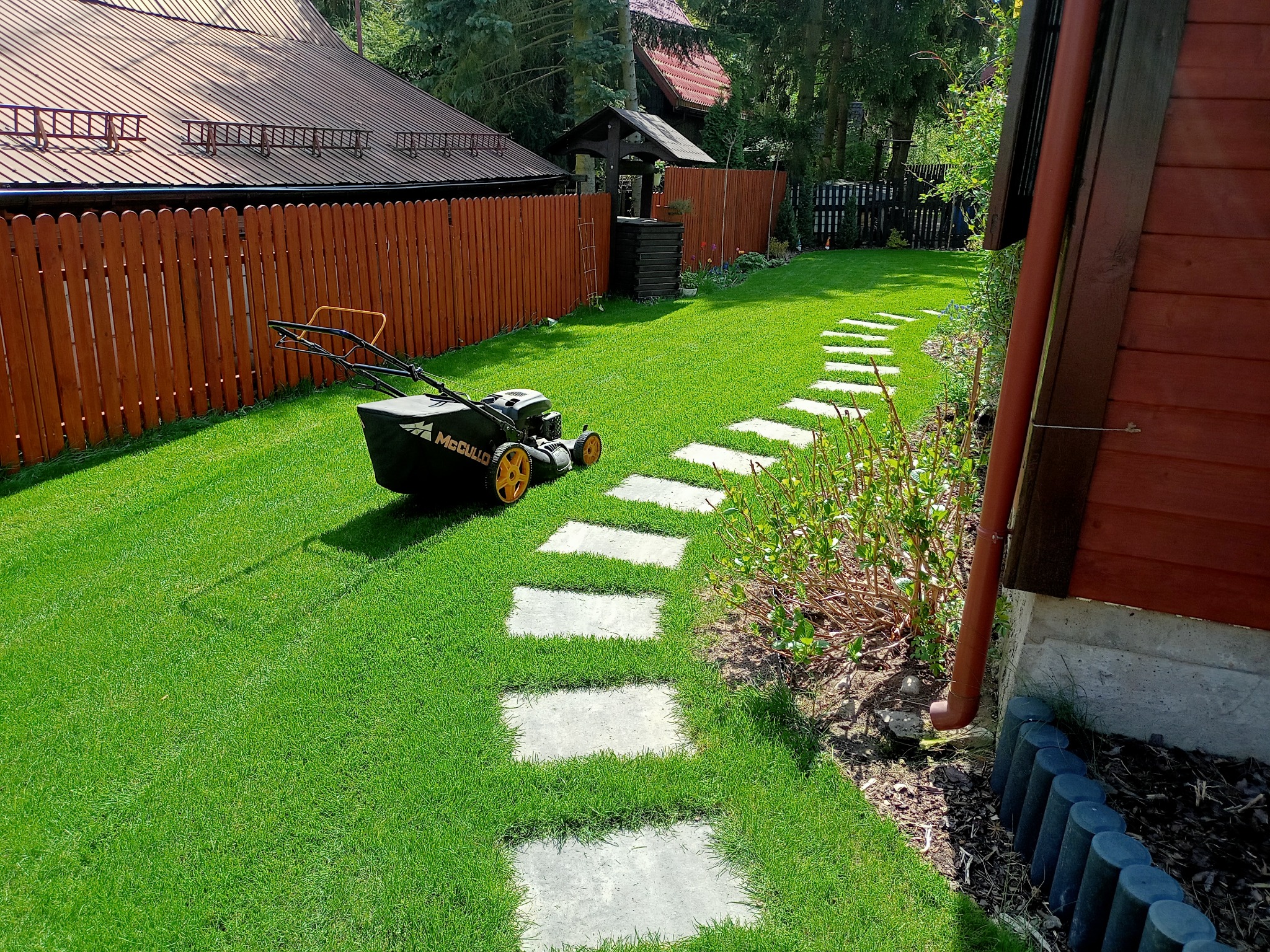 Kosiarka spalinowa McCulloch z żółtymi kołami na świeżo skoszonej, intensywnie zielonej trawie w ogrodzie, widoczna ścieżka z kamiennych płyt, drewniany płot i studnia w tle.