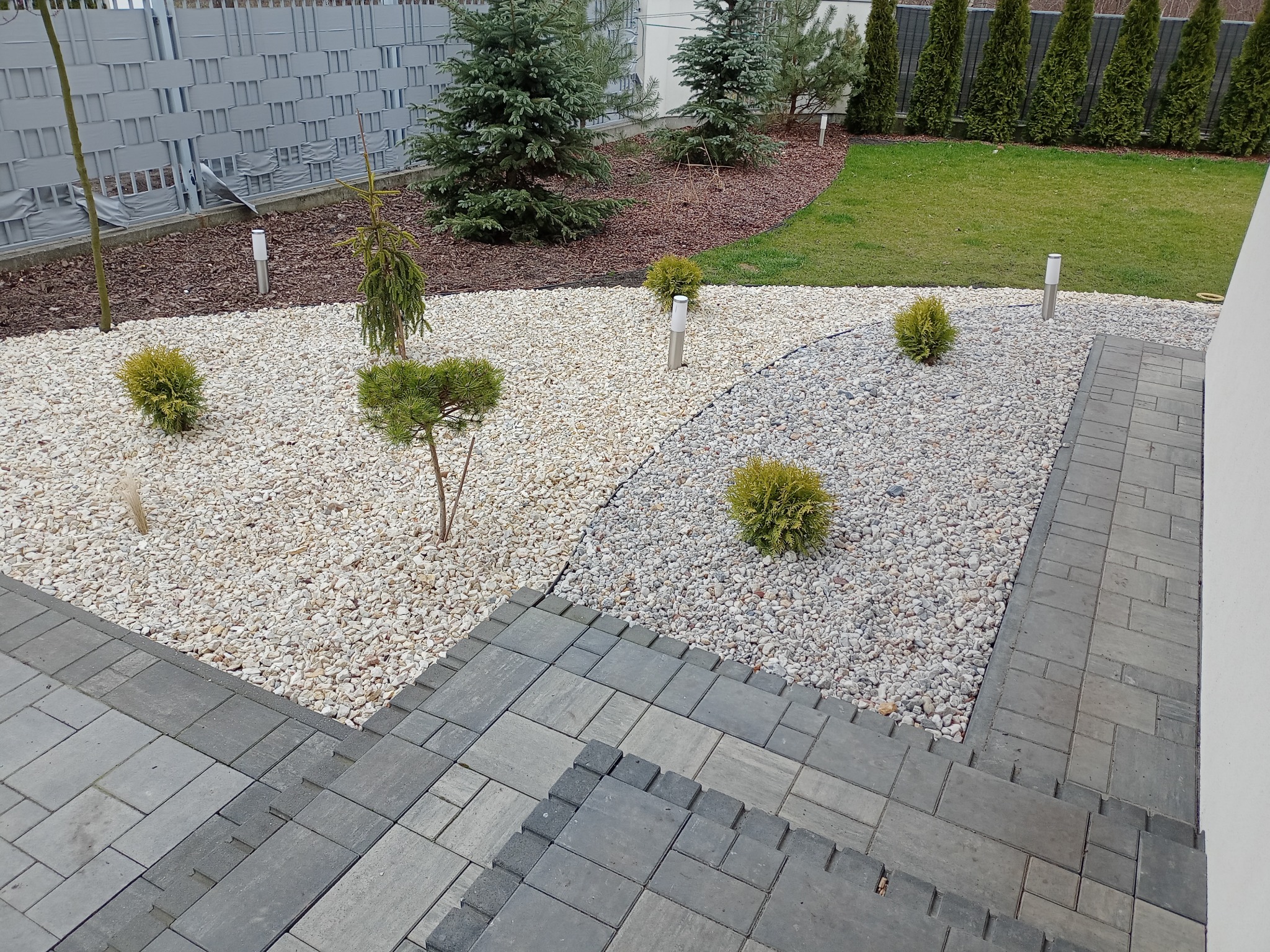 Ogród z jasnymi kamieniami, niskimi krzewami kulistymi i młodymi drzewami iglastymi, obrzeża z kostki brukowej, widok z góry na fragment ogrodu przy domu.