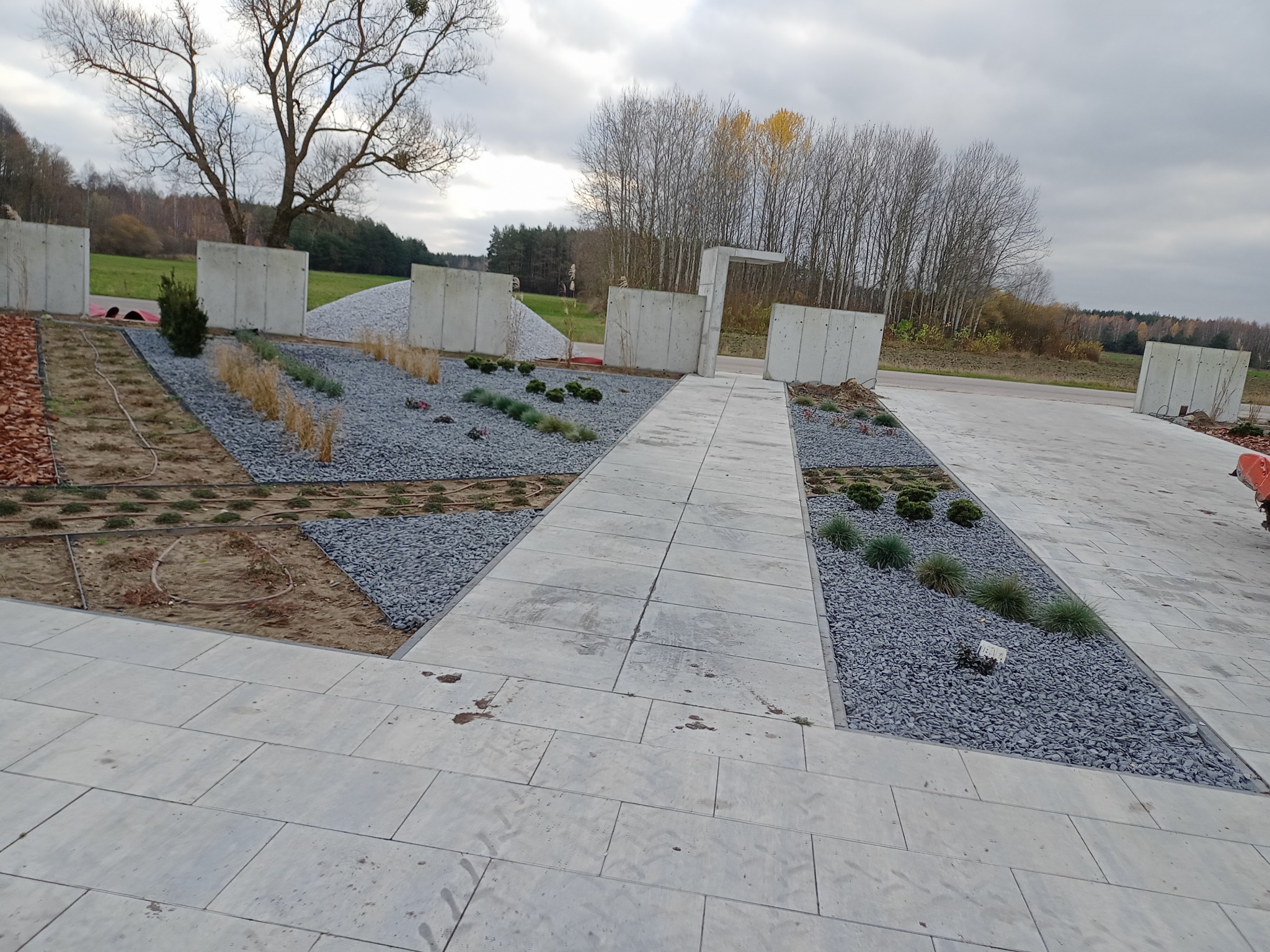 Nowoczesna aranżacja przestrzeni zewnętrznej z betonowymi płytami, szarym żwirem, ozdobnymi trawami i niskimi krzewami, otoczona polami i lasem pod pochmurnym niebem.