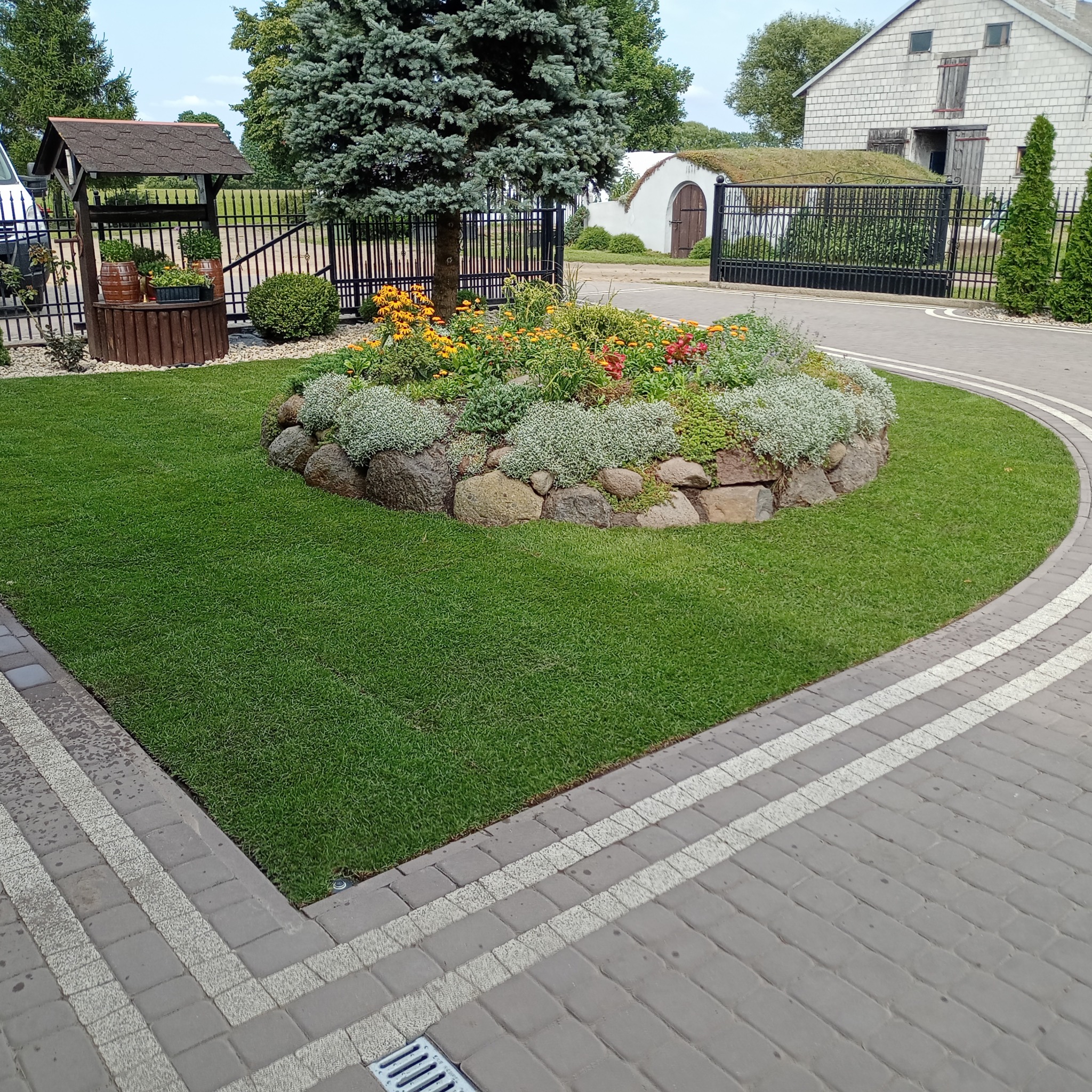 Ogród z równo przystrzyżonym trawnikiem, okrągłą rabatą kwiatową otoczoną kamieniami, studnią z drewnianą obudową i budynkiem z dachem pokrytym darnią w tle.