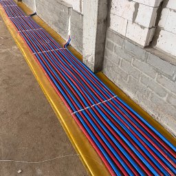 instal AP - Równolegle ułożone, czerwono-niebieskie rury ogrzewania podłogowego na żółtej folii izolacyjnej, przymocowane białymi opaskami, widok wzdłuż ściany z bloczków betonowych.