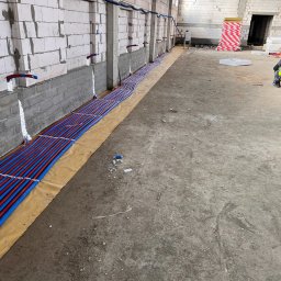 instal AP - Instalacja ogrzewania podłogowego w surowym stanie, widoczne rury grzewcze w kolorze niebieskim i czerwonym ułożone równolegle na folii izolacyjnej, z wyprowadzonymi przyłączami do ściany z betonu...