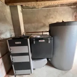 Nowoczesny piec na paliwo stałe z zasobnikiem i zbiornikiem akumulacyjnym w surowym wnętrzu, widoczne instalacje hydrauliczne i elektryczne.