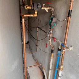 Złożona instalacja hydrauliczna z miedzianych i plastikowych rur, zaworów, pomp obiegowych oraz sterowników, zamontowana przy szarej ścianie w pomieszczeniu technicznym.