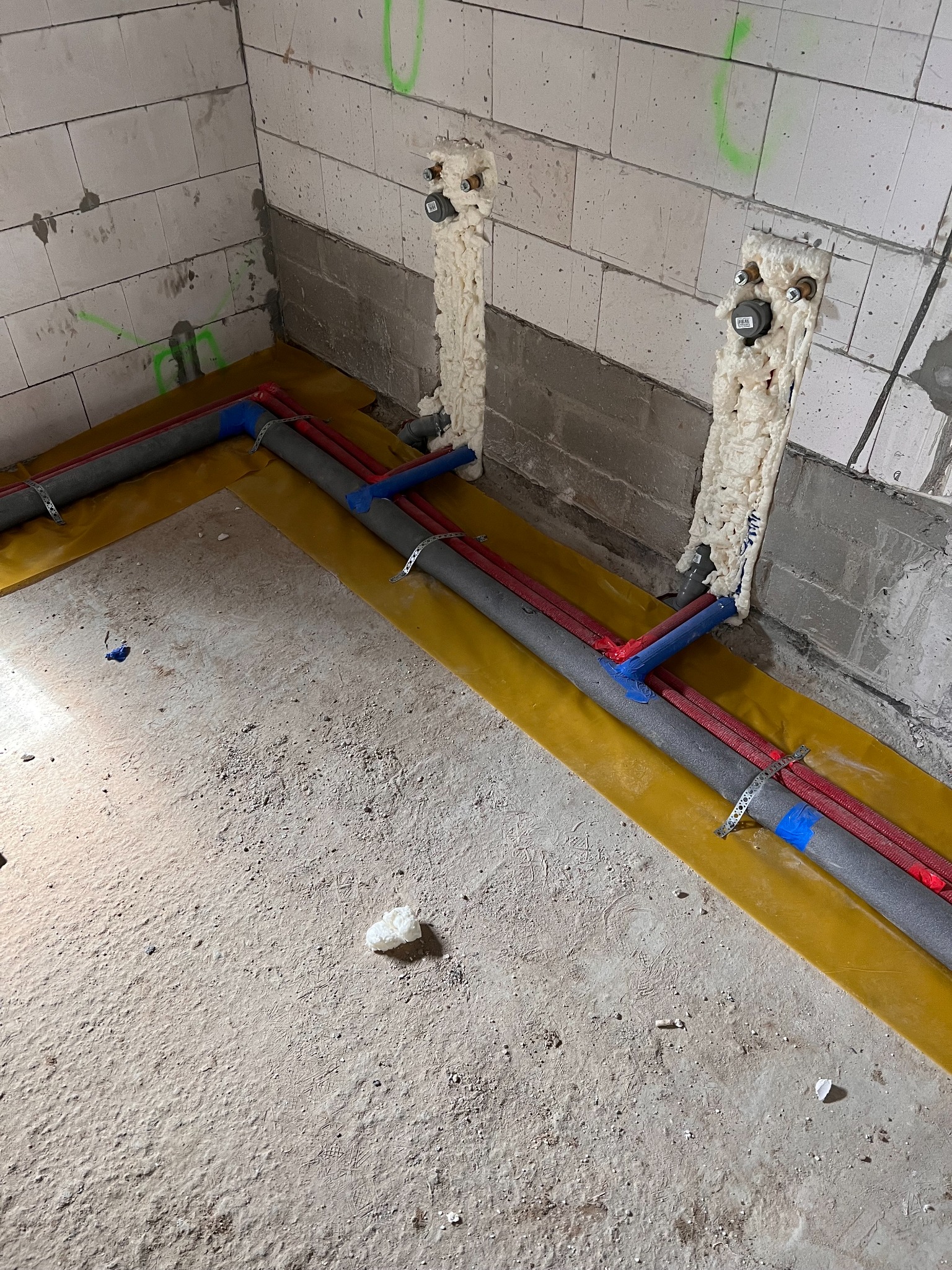 Montaż instalacji grzewczej podłogowej w surowym stanie: rury grzewcze w izolacji, mocowane do podłoża na żółtej folii, z pianką montażową wokół przyłączy ściennych.