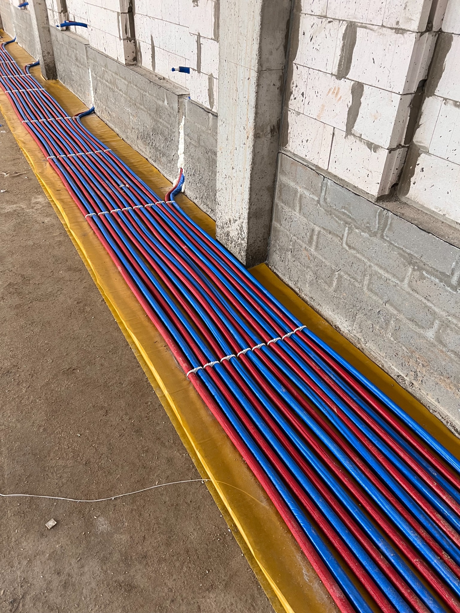 Równolegle ułożone, czerwono-niebieskie rury ogrzewania podłogowego na żółtej folii izolacyjnej, przymocowane białymi opaskami, widok wzdłuż ściany z bloczków betonowych.