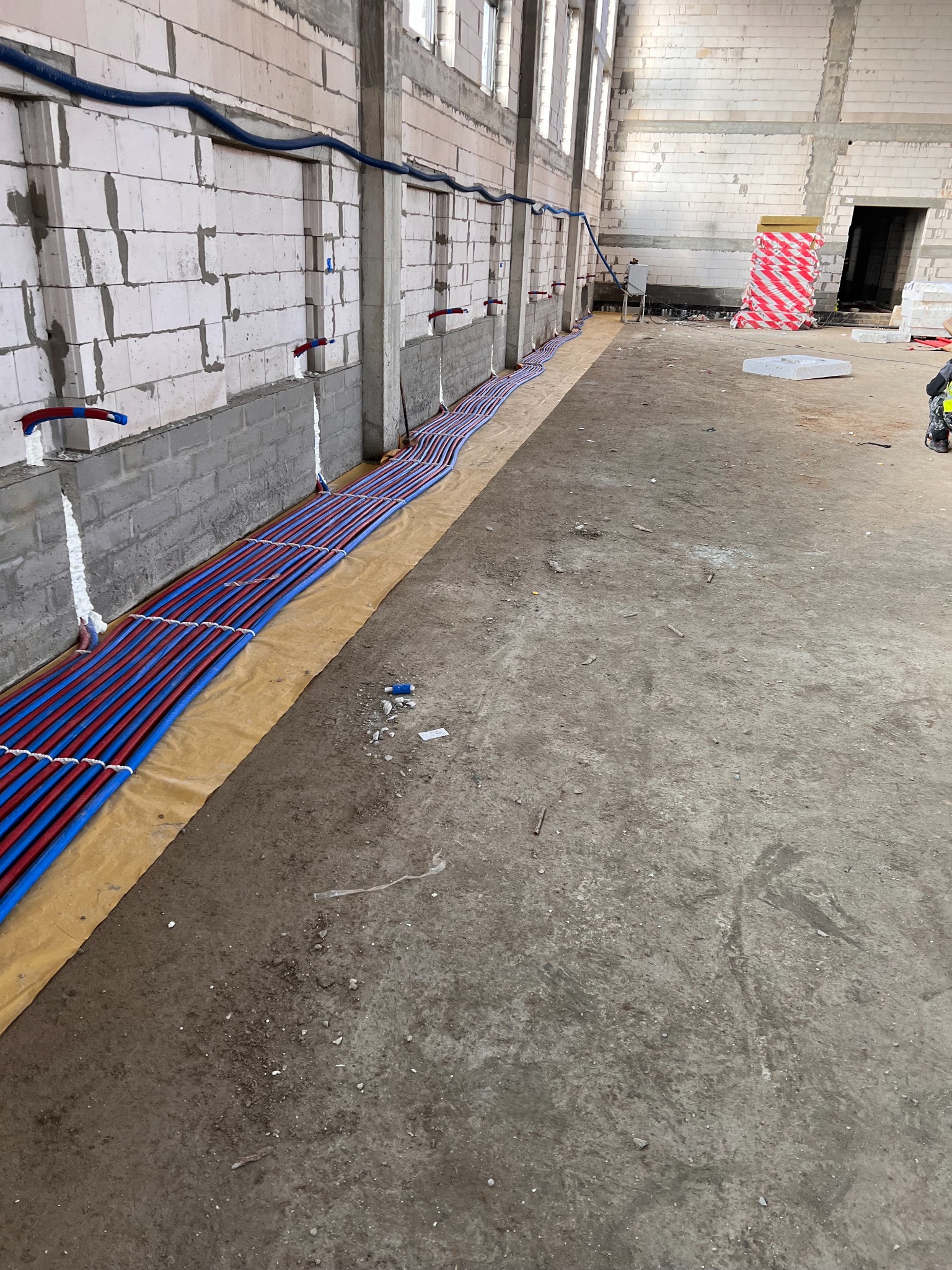 Instalacja ogrzewania podłogowego w surowym stanie, widoczne rury grzewcze w kolorze niebieskim i czerwonym ułożone równolegle na folii izolacyjnej, z wyprowadzonymi przyłączami do ściany z betonu...