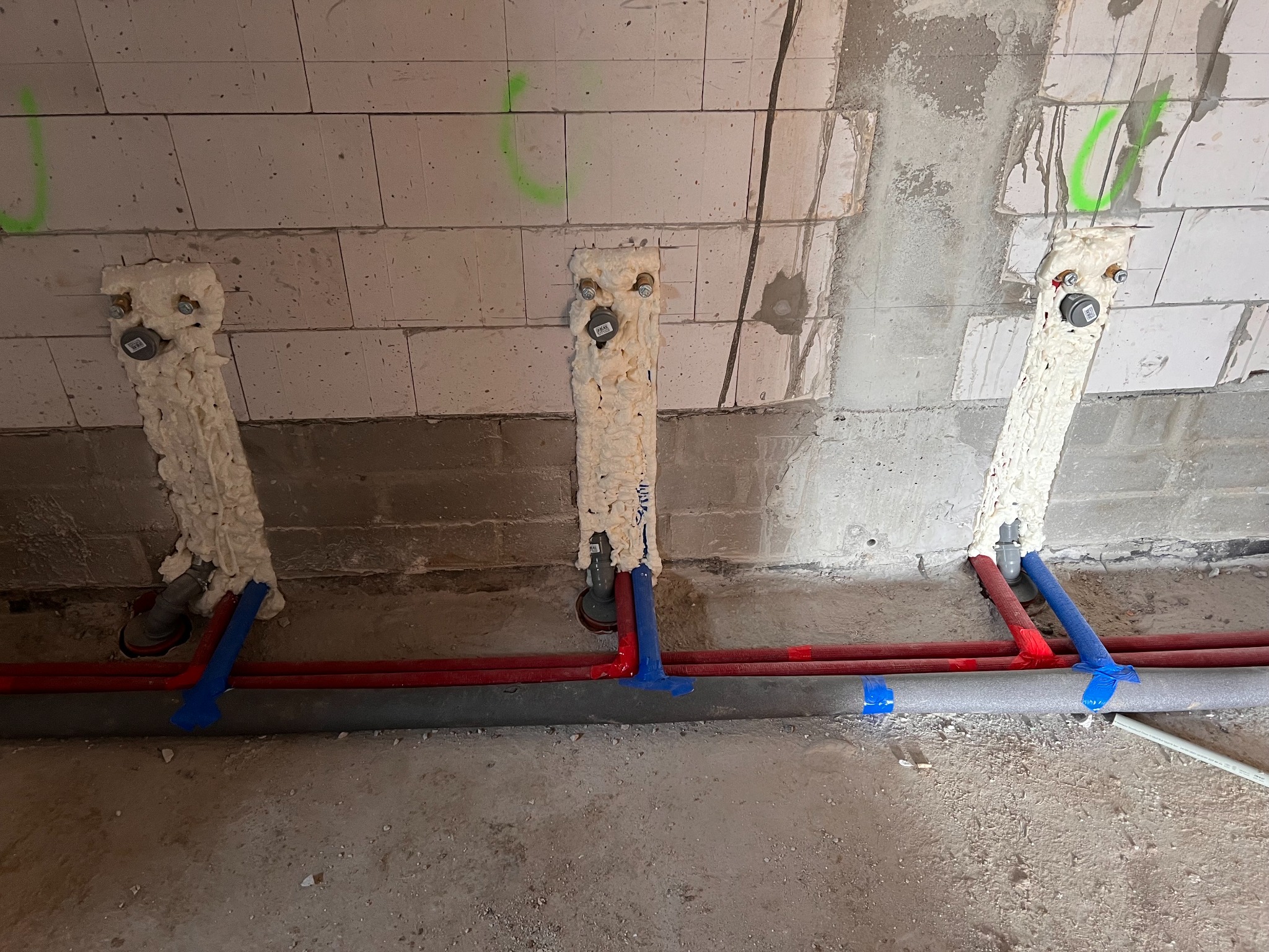 Świeżo zamontowane, zaizolowane pianką instalacje wodne z rurami czerwonymi i niebieskimi, przymocowane do ściany z betonu komórkowego z zielonymi oznaczeniami.