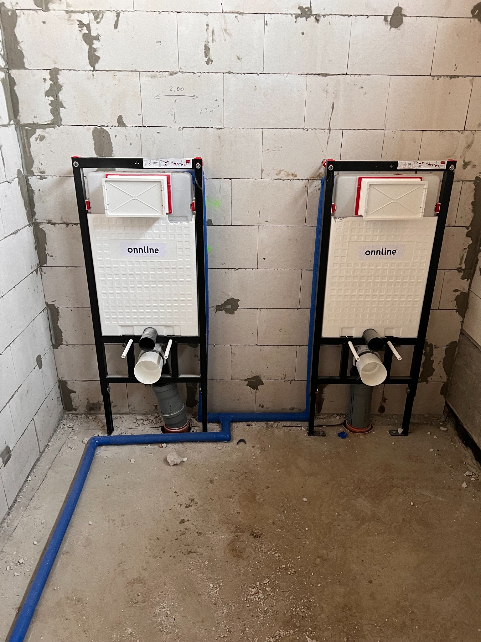 Dwa stelaże podtynkowe WC marki Onnline zamontowane na tle surowych ścian z betonu komórkowego, z widocznymi niebieskimi rurami doprowadzającymi wodę i szarą rurą odpływową.