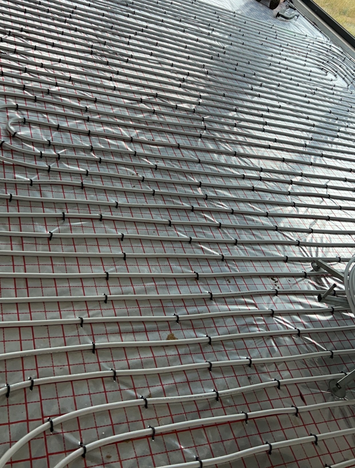 Białe rury ogrzewania podłogowego równolegle ułożone na folii z czerwoną siatką, przymocowane czarnymi uchwytami, widok z góry.