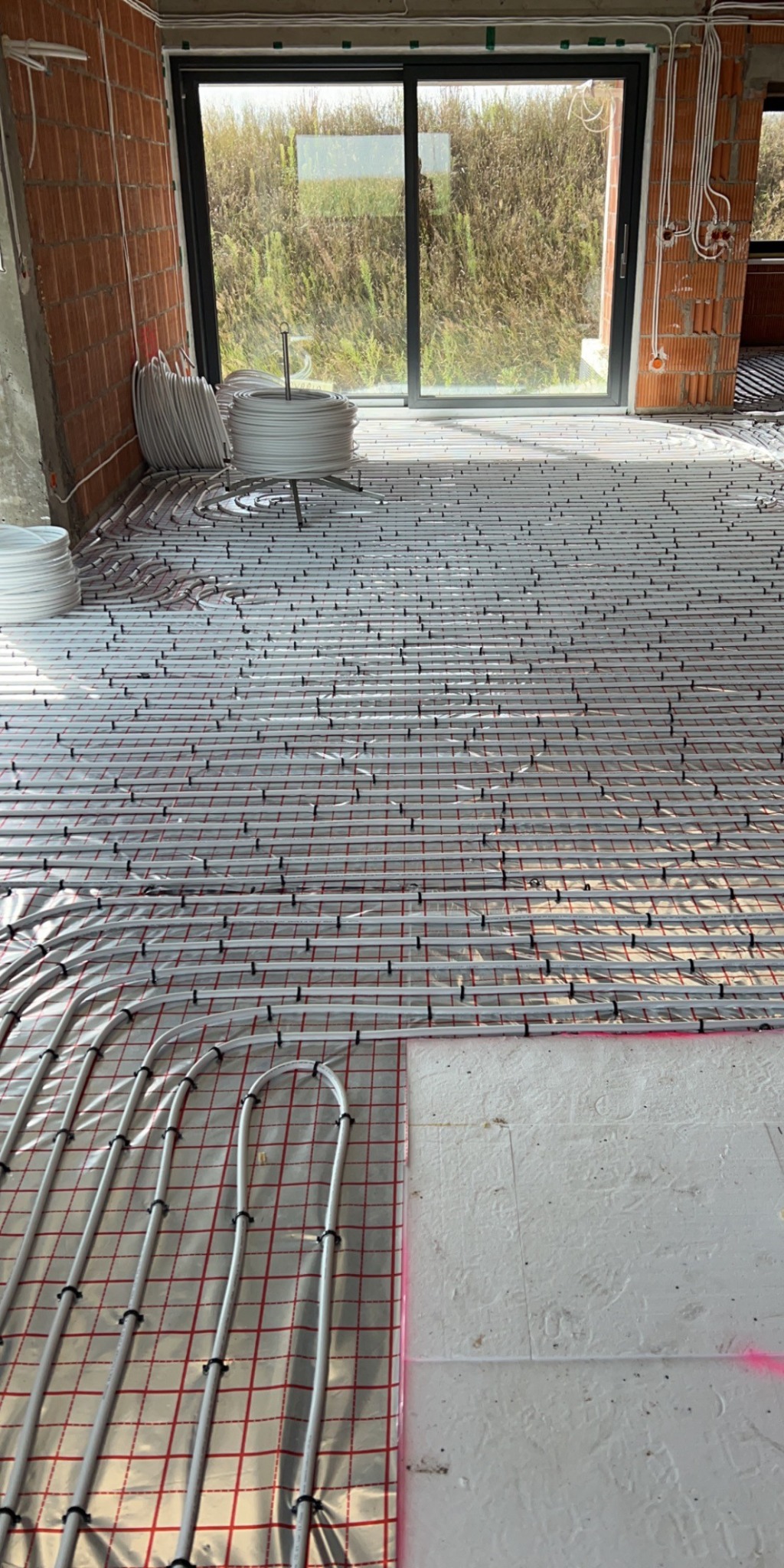 Rozłożona instalacja ogrzewania podłogowego na folii z rastrem, rury mocowane czarnymi klipsami, widoczne zwoje rur oraz okno z widokiem na zarośla.