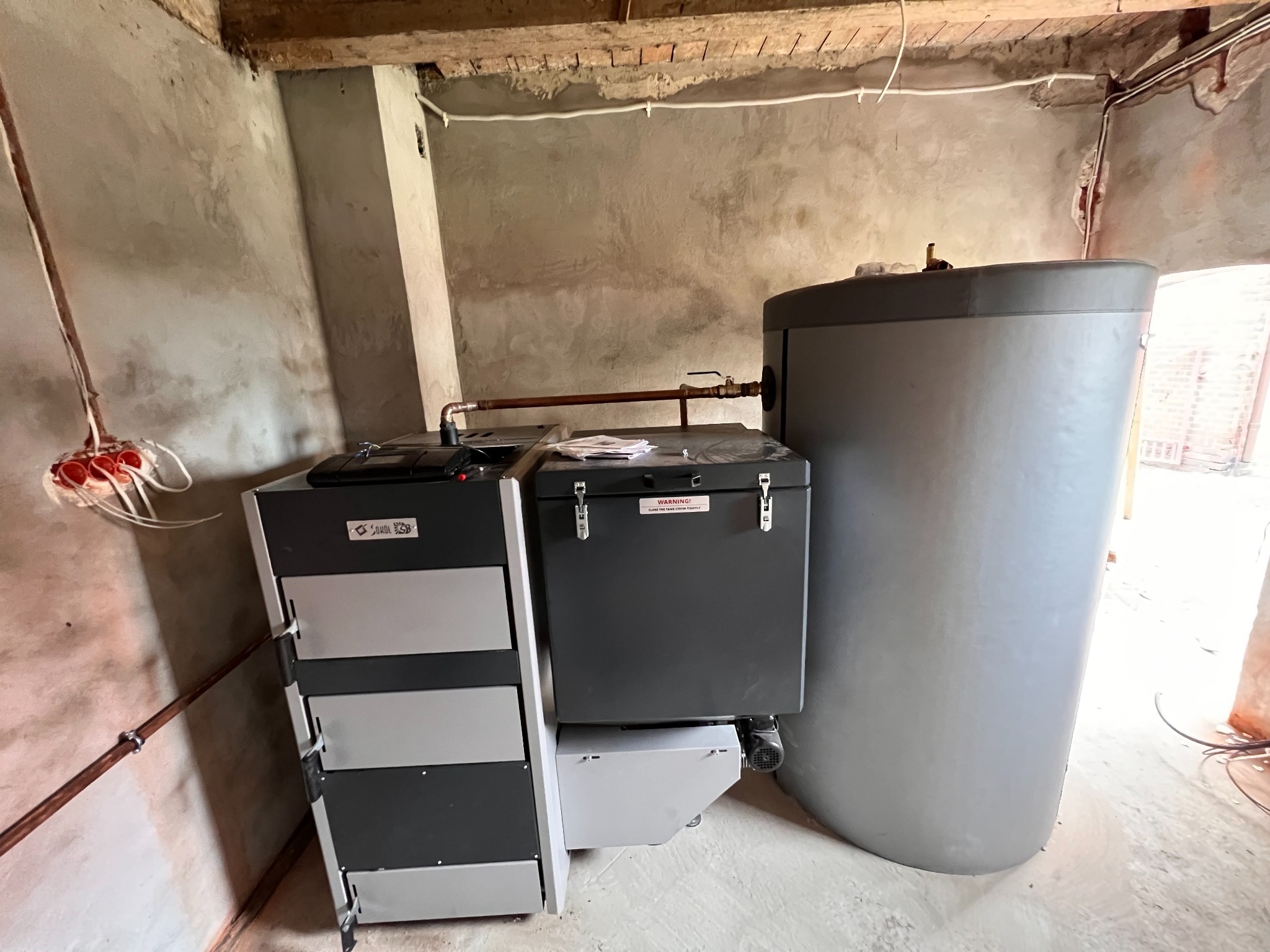 Nowoczesny piec na paliwo stałe z zasobnikiem i zbiornikiem akumulacyjnym w surowym wnętrzu, widoczne instalacje hydrauliczne i elektryczne.