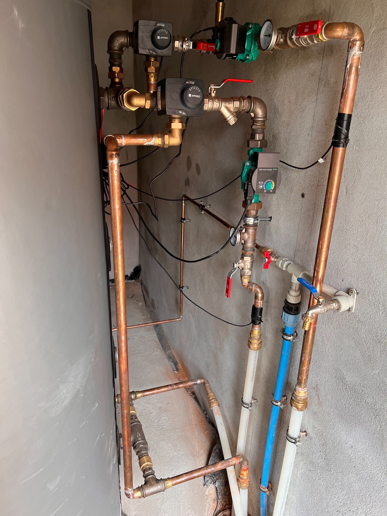 Złożona instalacja hydrauliczna z miedzianych i plastikowych rur, zaworów, pomp obiegowych oraz sterowników, zamontowana przy szarej ścianie w pomieszczeniu technicznym.