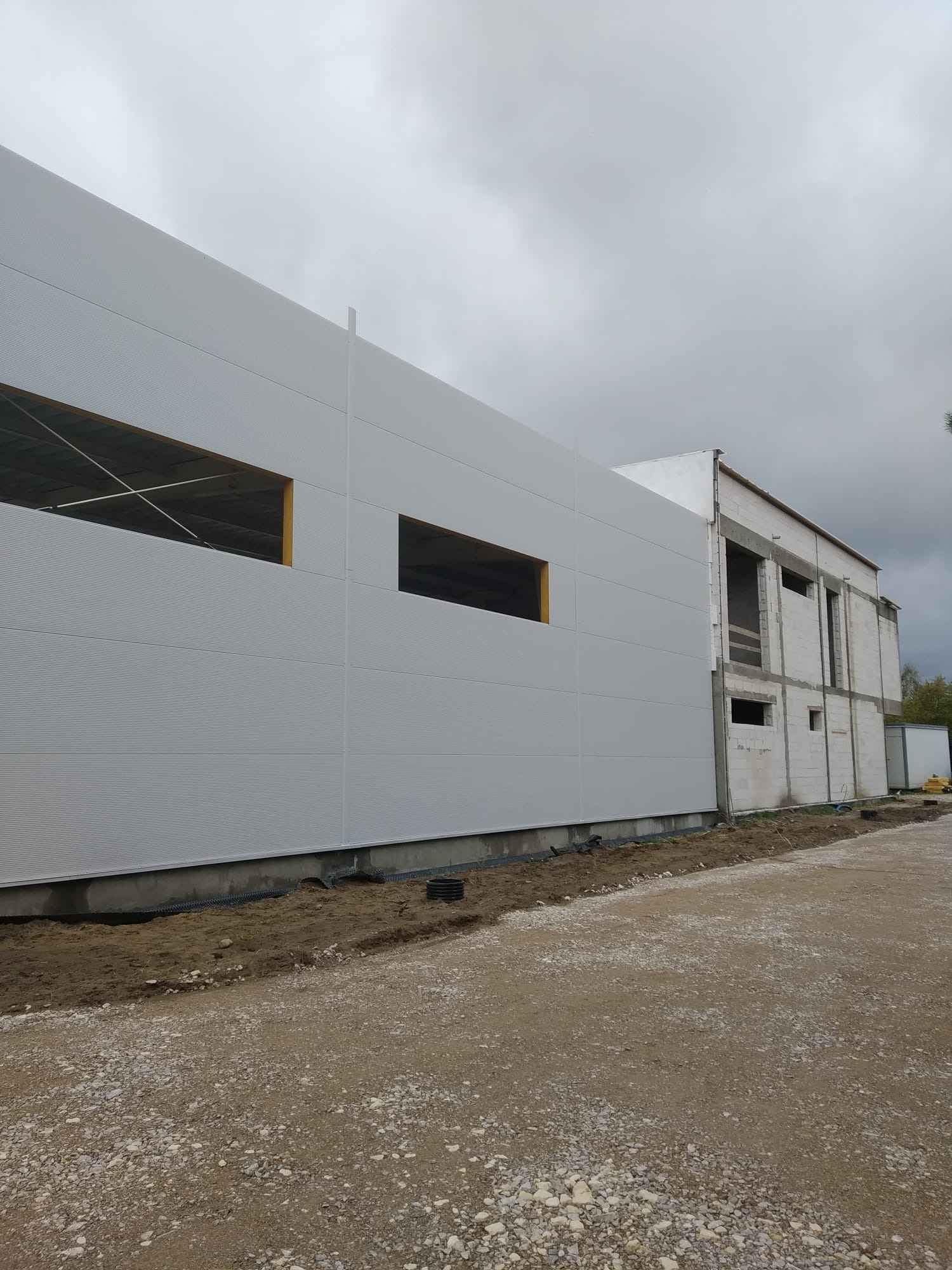 Budynek w trakcie budowy z szarymi panelami ściennymi i otworami okiennymi, widoczne elementy konstrukcyjne. Część budynku w surowym stanie, cegła i beton. Na pierwszym planie teren budowy z gruzem.