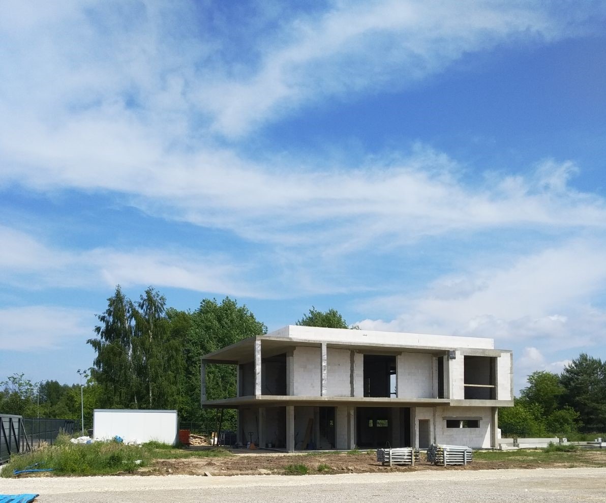 Surowy, dwukondygnacyjny budynek żelbetowy w trakcie budowy, widoczny szkielet konstrukcji na tle błękitnego nieba z chmurami i zieleni drzew.