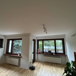 ACREO INTERNATIONAL SPÓŁKA Z OGRANICZONĄ ODPOWIEDZIALNOŚCIĄ - Jasne wnętrze po odświeżeniu: parkiet w odcieniach beżu, białe ściany z grzejnikami pod oknami w ciemnych ramach, czarny robot sprzątający przy ścianie.