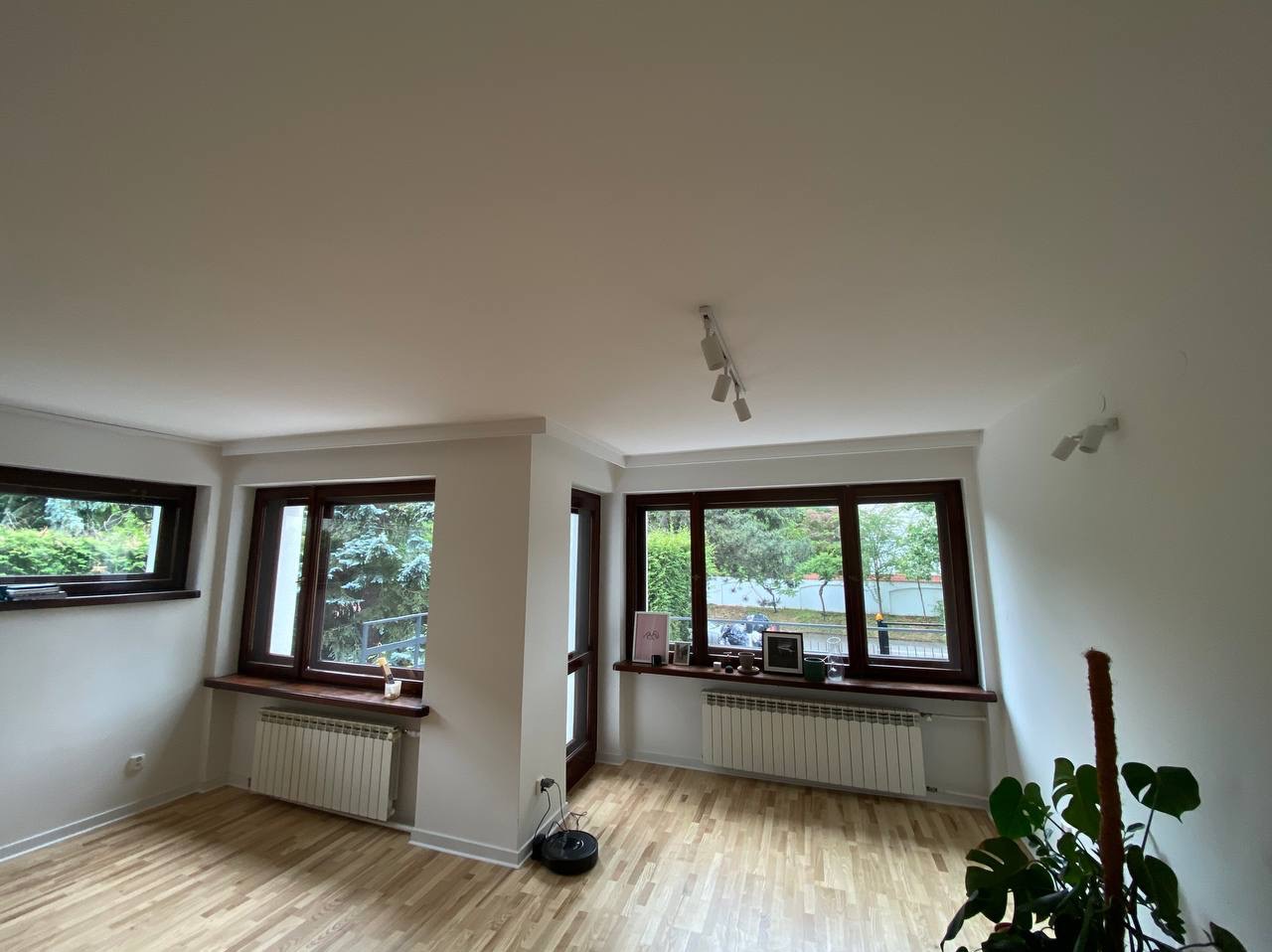 Jasne wnętrze po odświeżeniu: parkiet w odcieniach beżu, białe ściany z grzejnikami pod oknami w ciemnych ramach, czarny robot sprzątający przy ścianie.