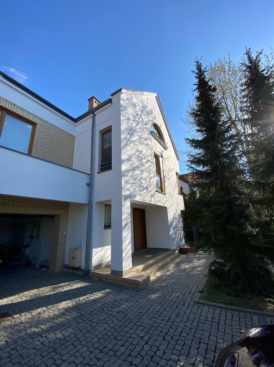 Nowoczesny dom z białymi ścianami i ceglanymi elementami, widok z dołu w słoneczny dzień, cień drzew na fasadzie, kostka brukowa na podjeździe.