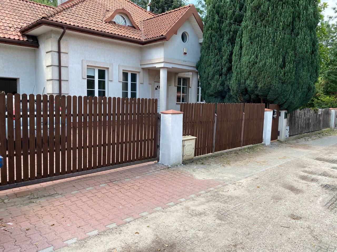 Brązowe, drewniane ogrodzenie posesji z białymi słupkami, widok z zewnątrz na dom z jasną elewacją i czerwoną dachówką.