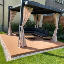 Roman Dąbkowski - Nowoczesna pergola z zasłonami na kompozytowym tarasie, otoczona zielenią. Minimalistyczny design, drewniany stół i sofa z poduszkami.
