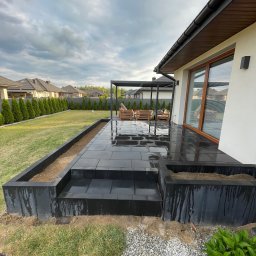 Roman Dąbkowski - Nowoczesny taras z czarnej kostki brukowej, z podwyższonymi rabatami, pergola i meble ogrodowe w tle, po deszczu, widok na dom i ogród.