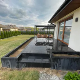 Nowoczesny taras z czarnej kostki brukowej, z podwyższonymi rabatami, pergola i meble ogrodowe w tle, po deszczu, widok na dom i ogród.