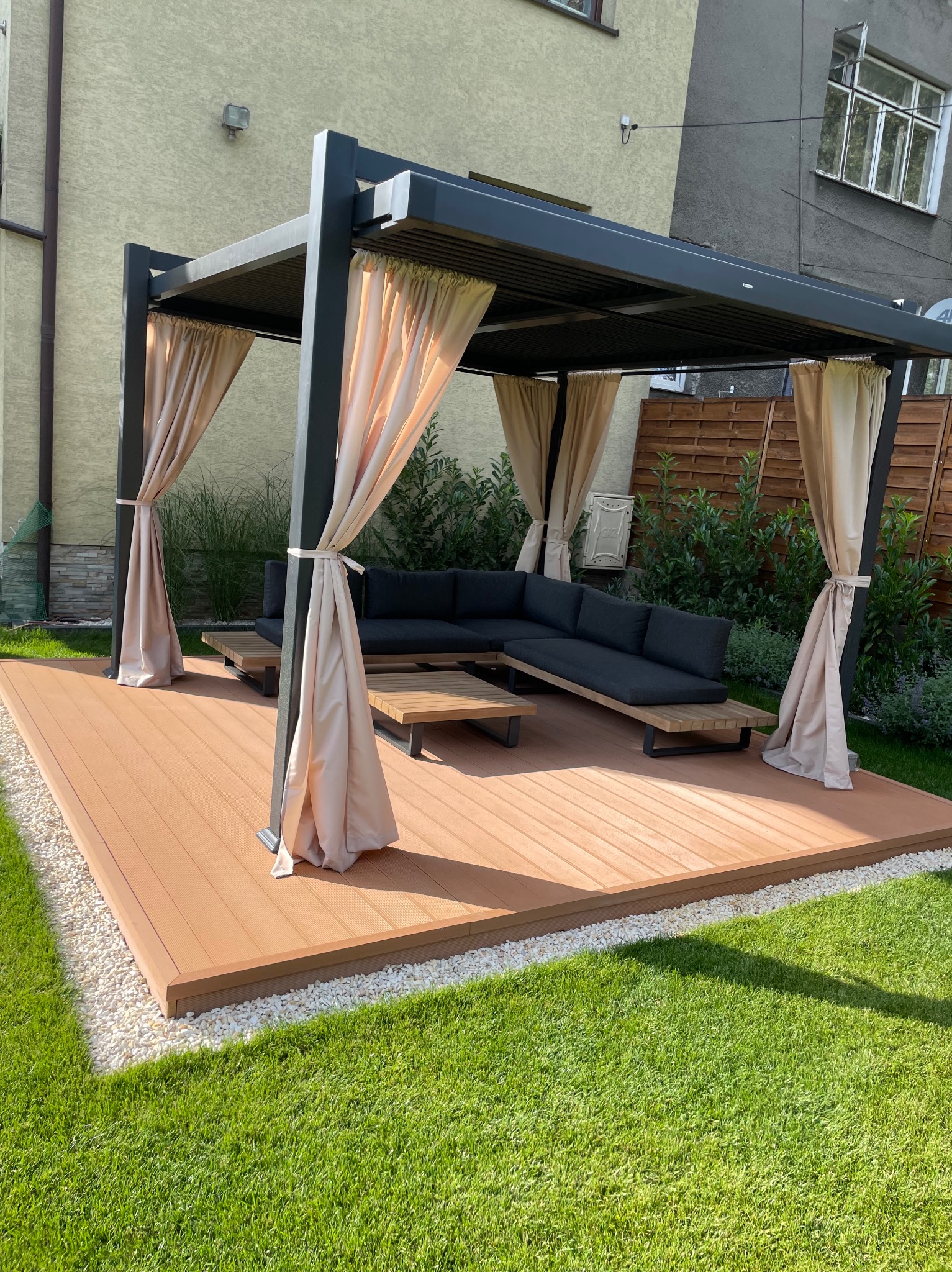 Nowoczesna pergola z zasłonami na kompozytowym tarasie, otoczona zielenią. Minimalistyczny design, drewniany stół i sofa z poduszkami.