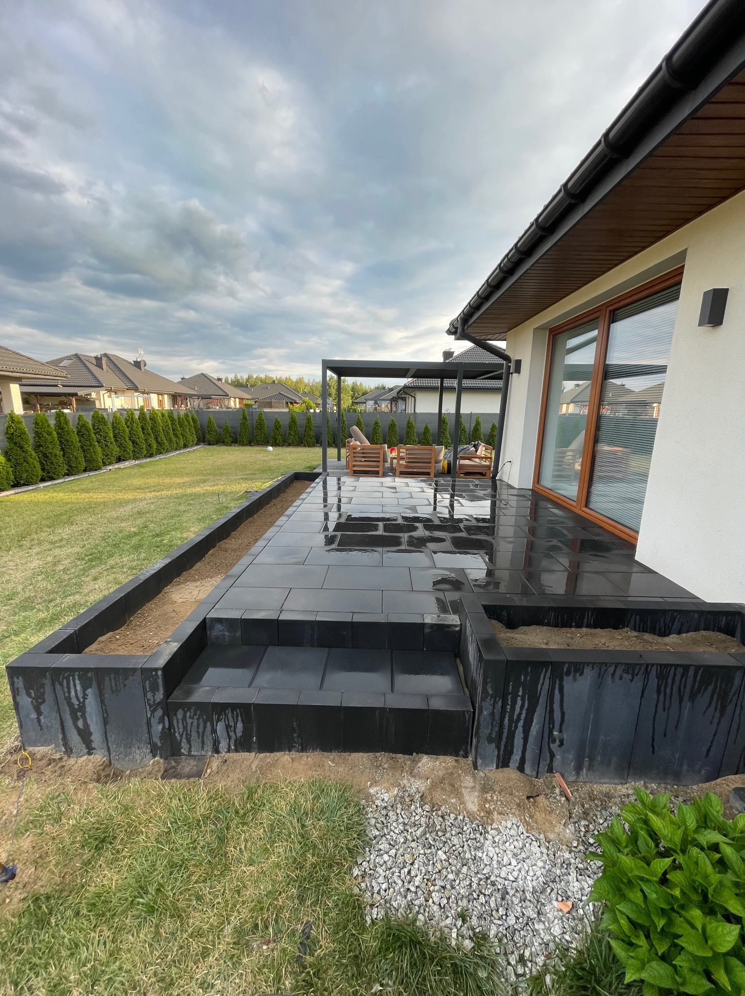 Nowoczesny taras z czarnej kostki brukowej, z podwyższonymi rabatami, pergola i meble ogrodowe w tle, po deszczu, widok na dom i ogród.