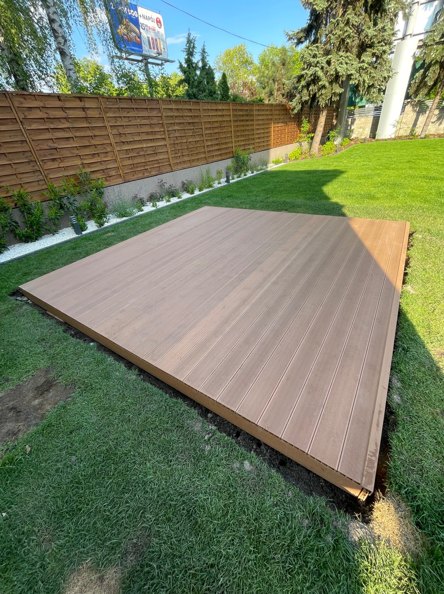 Nowoczesny taras kompozytowy w ogrodzie, widok z góry. Brązowe deski ułożone na trawie, w tle drewniany płot i zieleń. Estetyczne wykończenie.