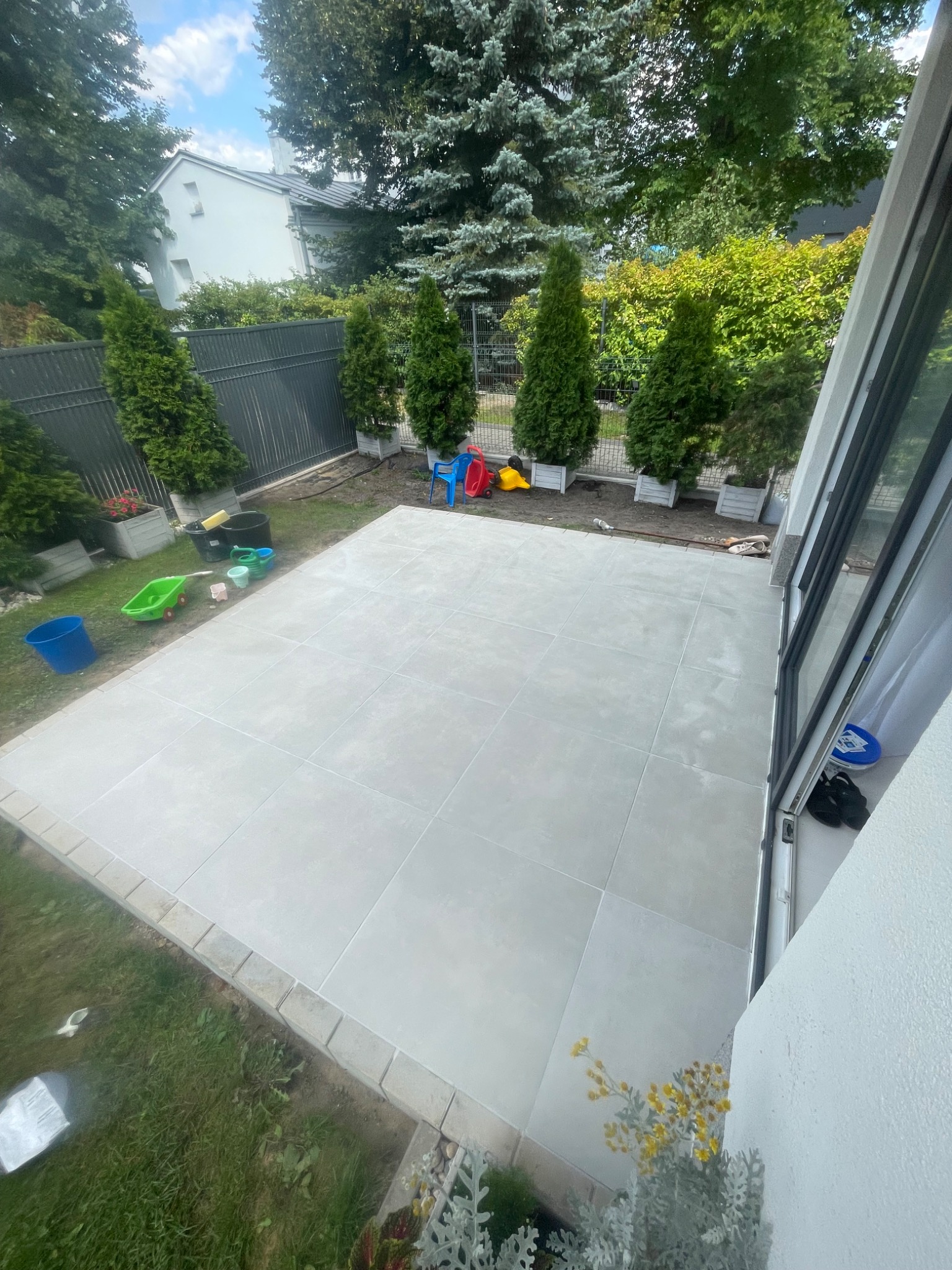 Wykończony taras z jasnych, kwadratowych płyt w ogrodzie z tujami. Widoczne zabawki dziecięce i narzędzia ogrodnicze. Ujęcie z wnętrza domu przez otwarte okno.