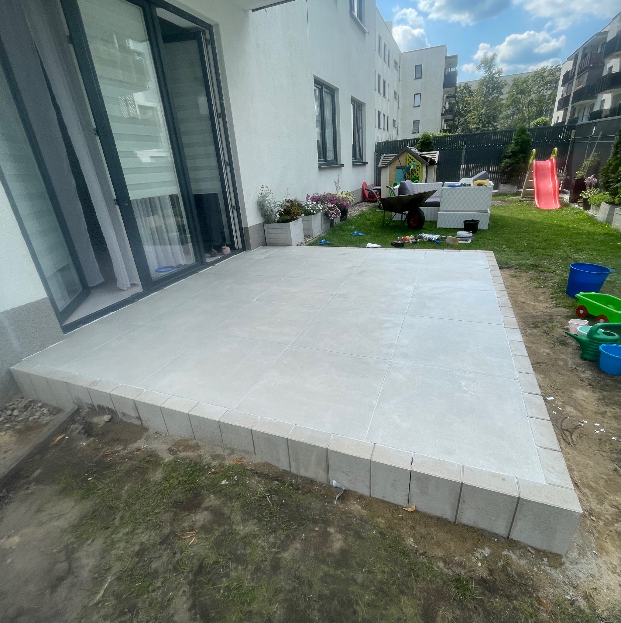 Jasny taras z płytek na podwyższeniu, widok z góry, z elementami ogrodu i domu w tle, w słoneczny dzień. Widoczne fugi i obrzeża tarasu.