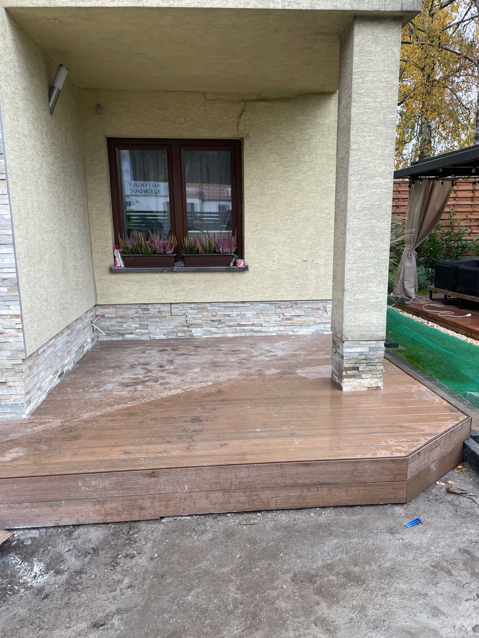 Wykończony taras z desek w odcieniach brązu, przylegający do budynku z oknem i kwiatami. Widoczny fragment ogrodu z zadaszeniem i meblami.