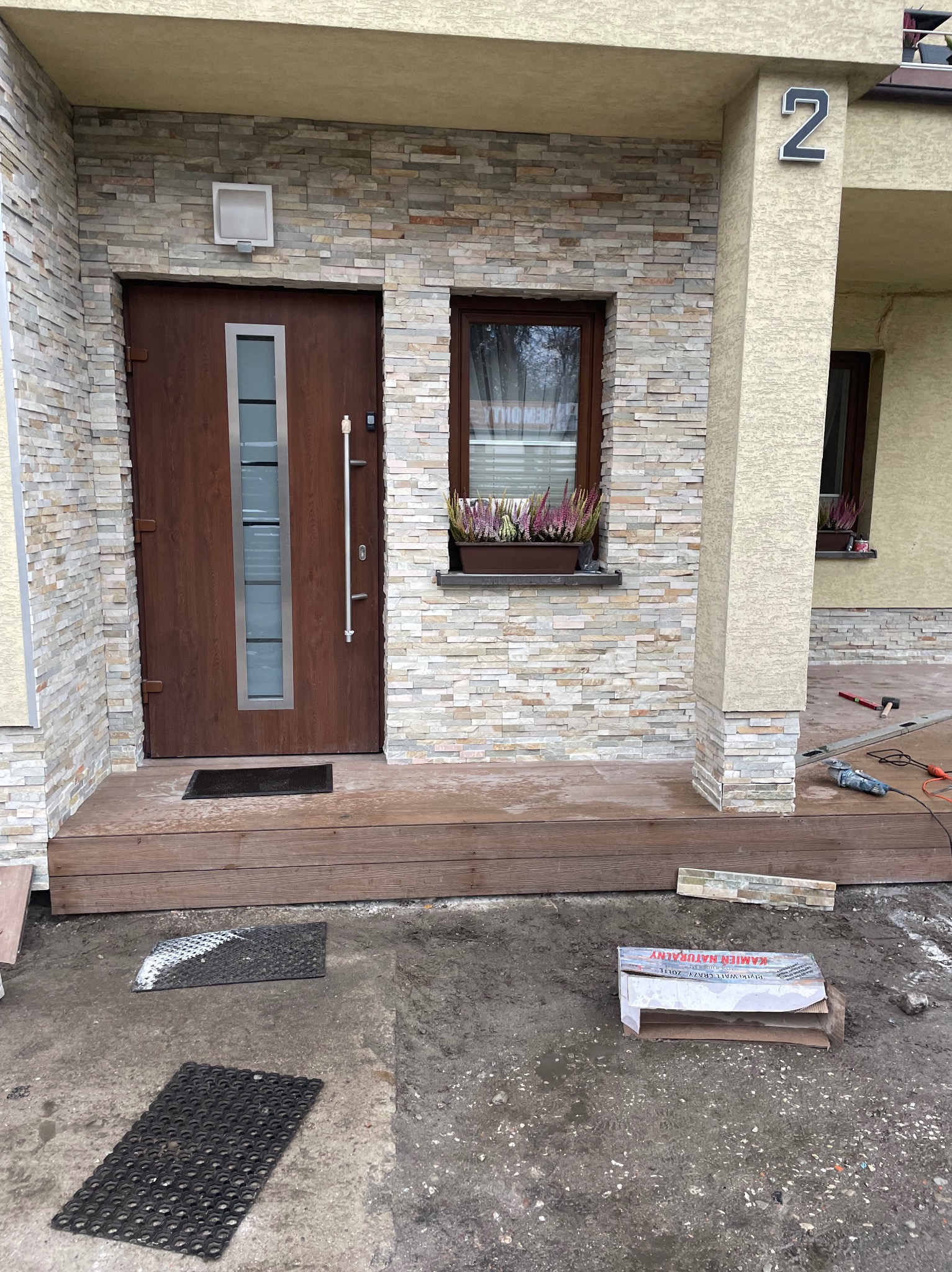 Montaż elewacji z kamienia dekoracyjnego przy wejściu do domu. Widoczne narzędzia i materiały budowlane. Etap prac wykończeniowych.