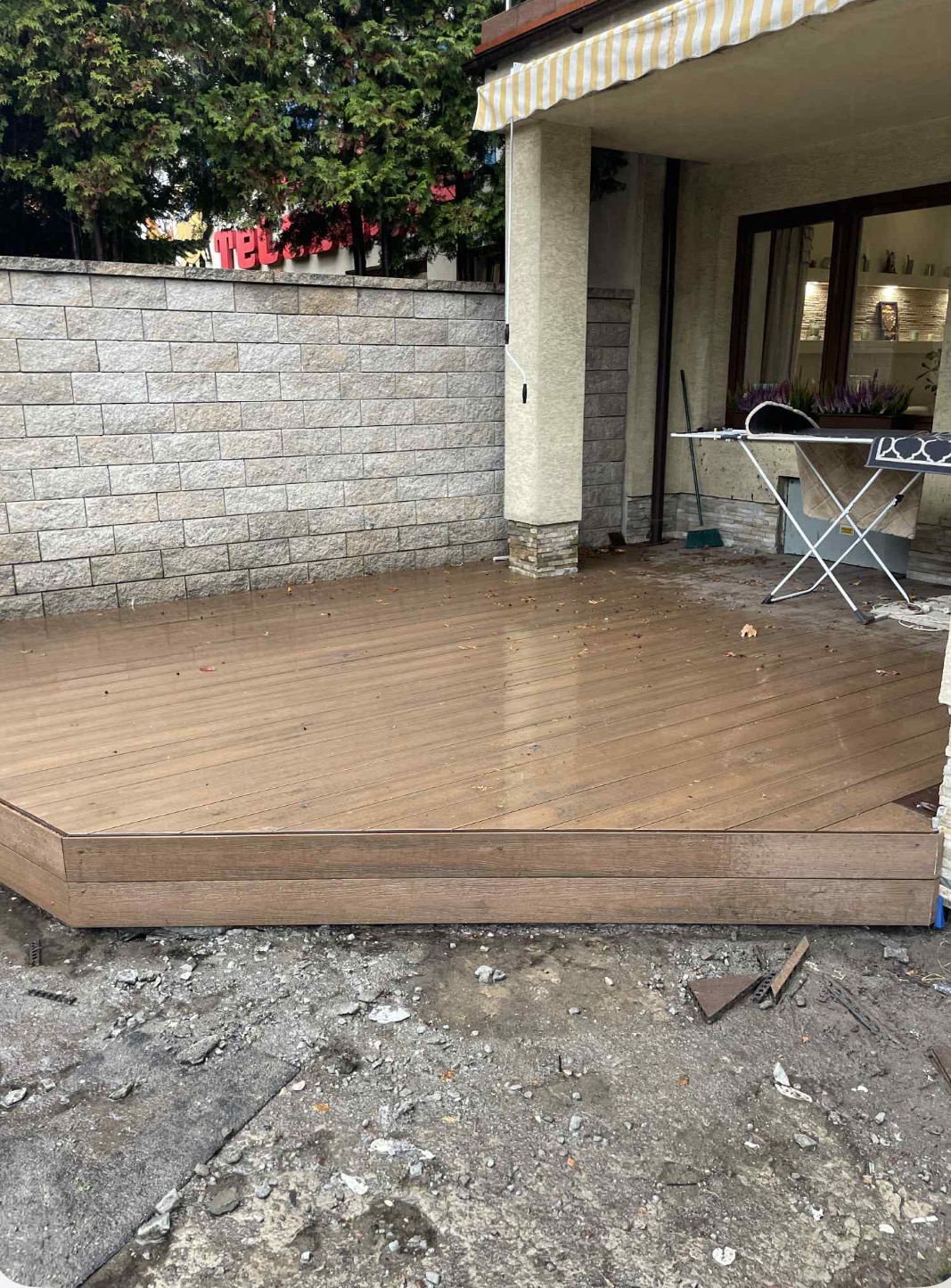 Wykończony taras kompozytowy z widoczną strukturą desek, przylegający do budynku z elementami kamiennej elewacji i fragmentem ogrodu w tle.