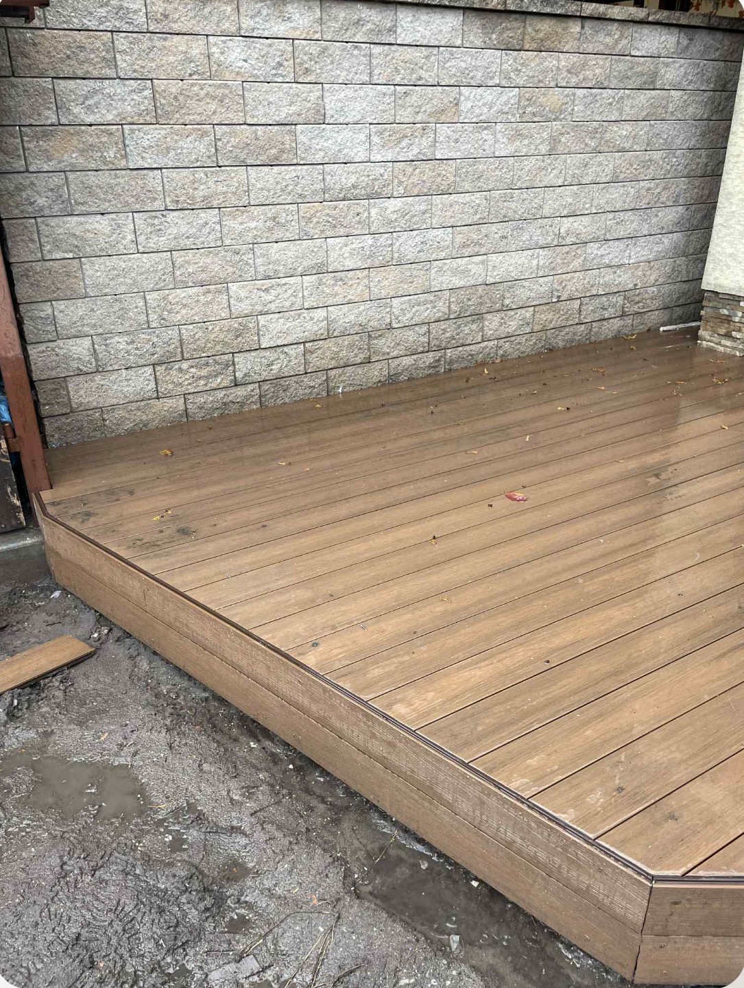 Kątowe ujęcie tarasu z desek kompozytowych w odcieniach brązu, z widoczną fakturą drewna i szarą ścianą z cegły w tle. Na deskach leżą pojedyncze liście.