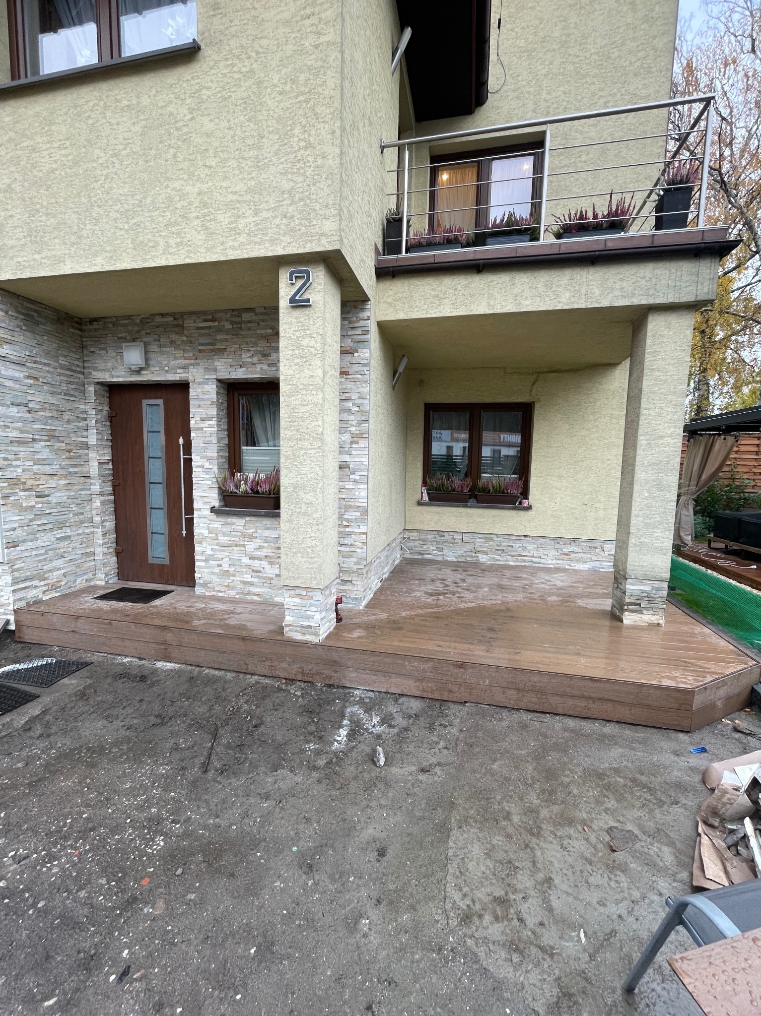 Nowoczesny dom z drewnianym tarasem i balkonem. Elewacja z kamienia i jasnego tynku. Okna z kwiatami. Numer domu '2'.