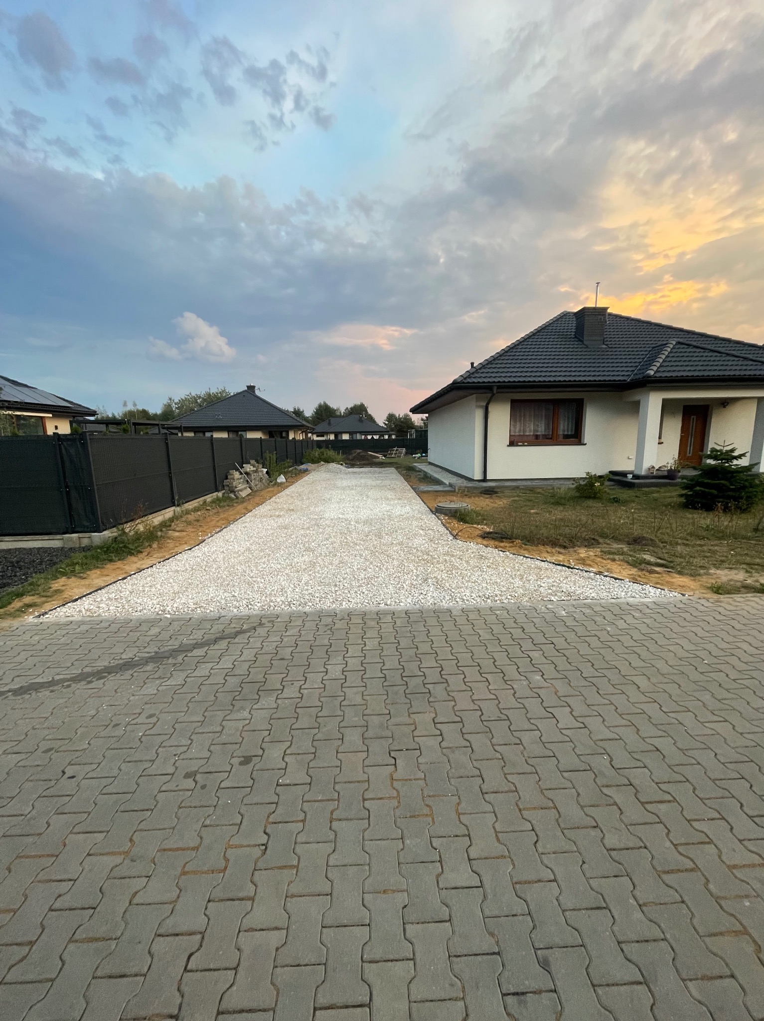 Ułożona kostka brukowa prowadząca do domu z białym kamieniem po bokach, ogrodzenie z siatki po lewej stronie, wieczorne niebo w tle.